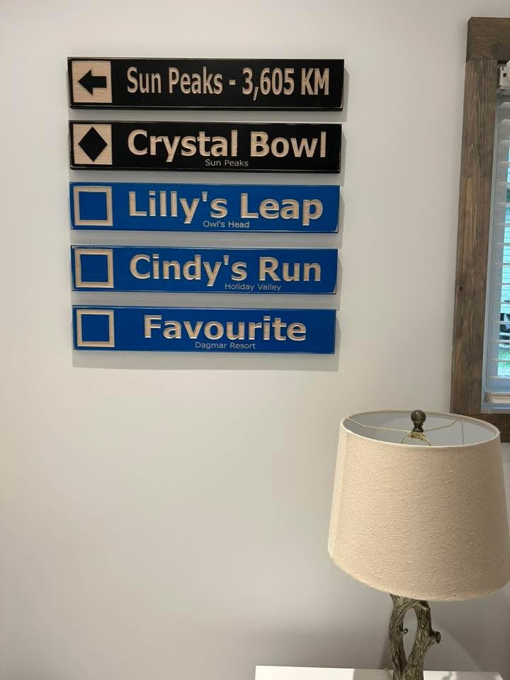 Wall-mounted custom wood ski trail signs with resort names above a rustic lamp.