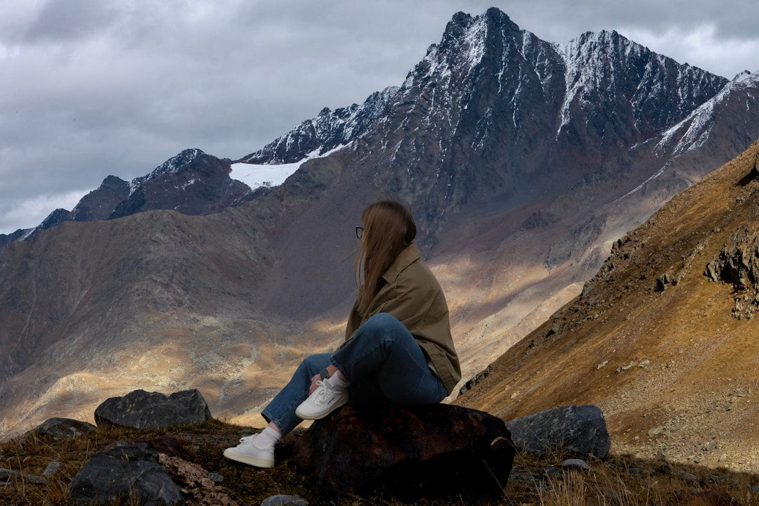 Woman sitting on a rock overlooking rugged mountains with snowy peaks and cloudy sky