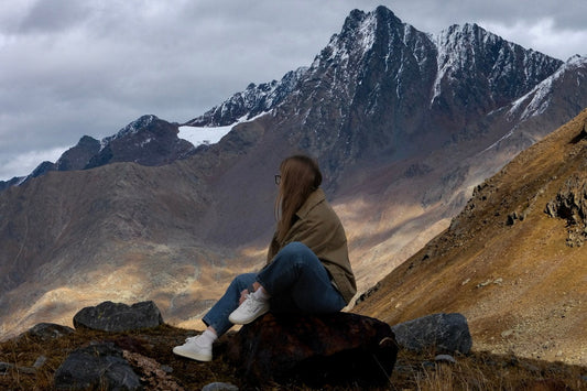 Woman sitting on a rock overlooking rugged mountains with snowy peaks and cloudy sky