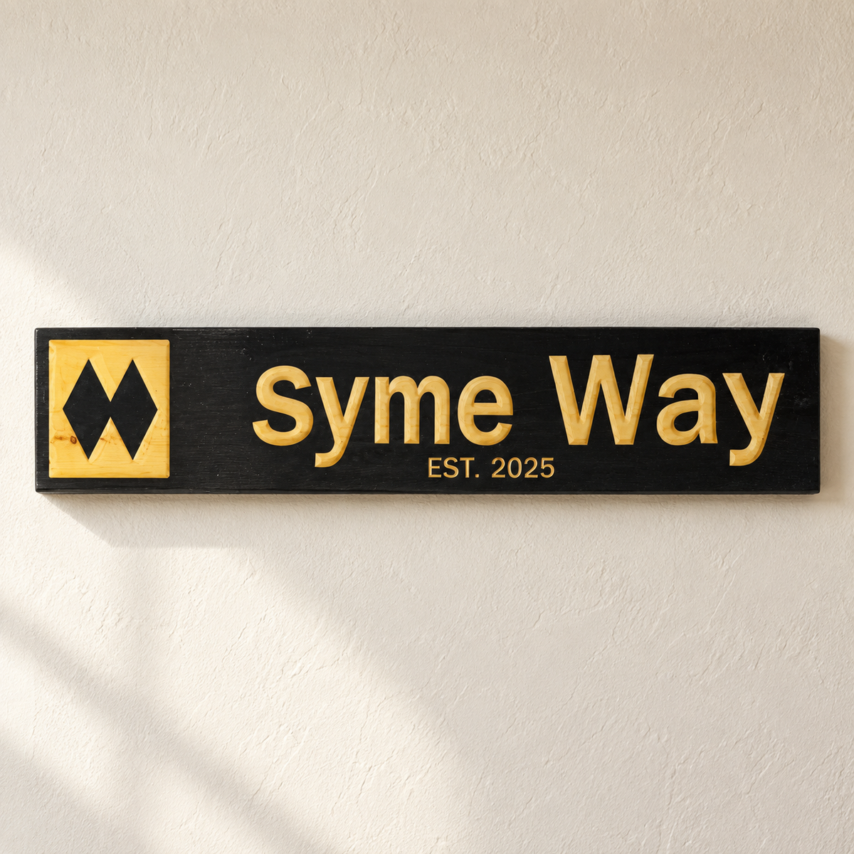 A rectangular black wooden sign mounted on a textured off-white wall. The sign features raised gold lettering that reads 'Syme Way' with 'EST.