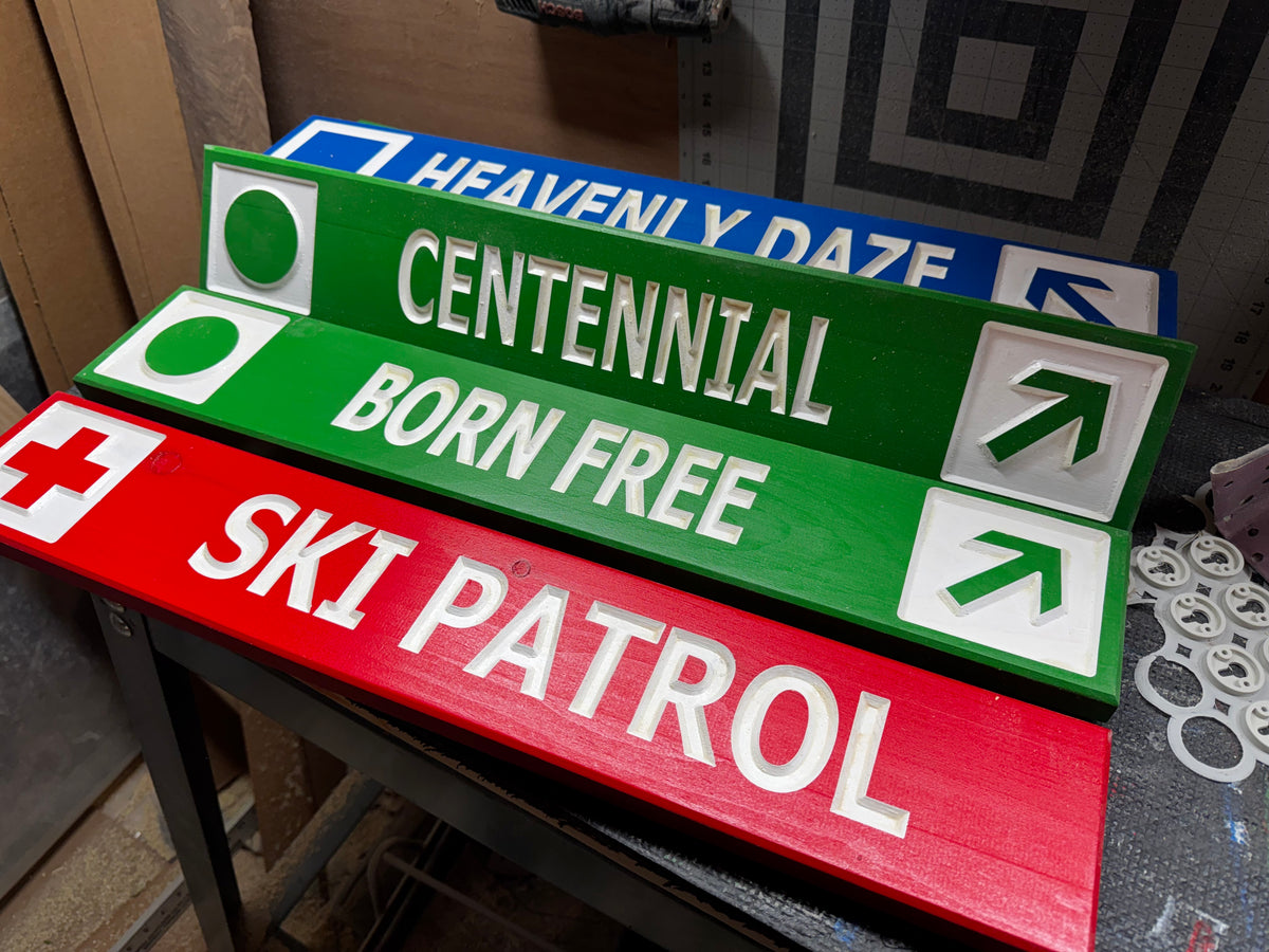 Four painted wooden signboards with directional text and symbols, including green 'CENTENNIAL BORN FREE' and 'HEAVENLY DAZE' signs and a red 'SKI PATROL' sign, displayed on a workbench.
