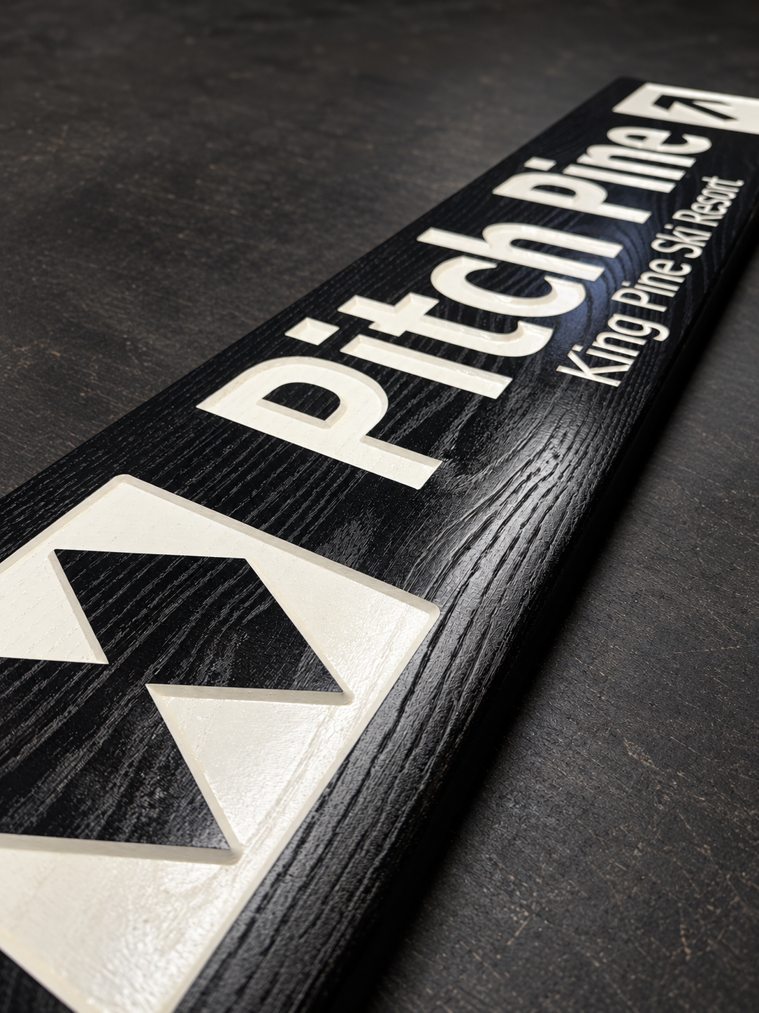 A close-up diagonal shot of a black wooden sign with white lettering reading 'Pitch Pine Ski Resort' and a large white geometric logo, placed on a dark textured surface.