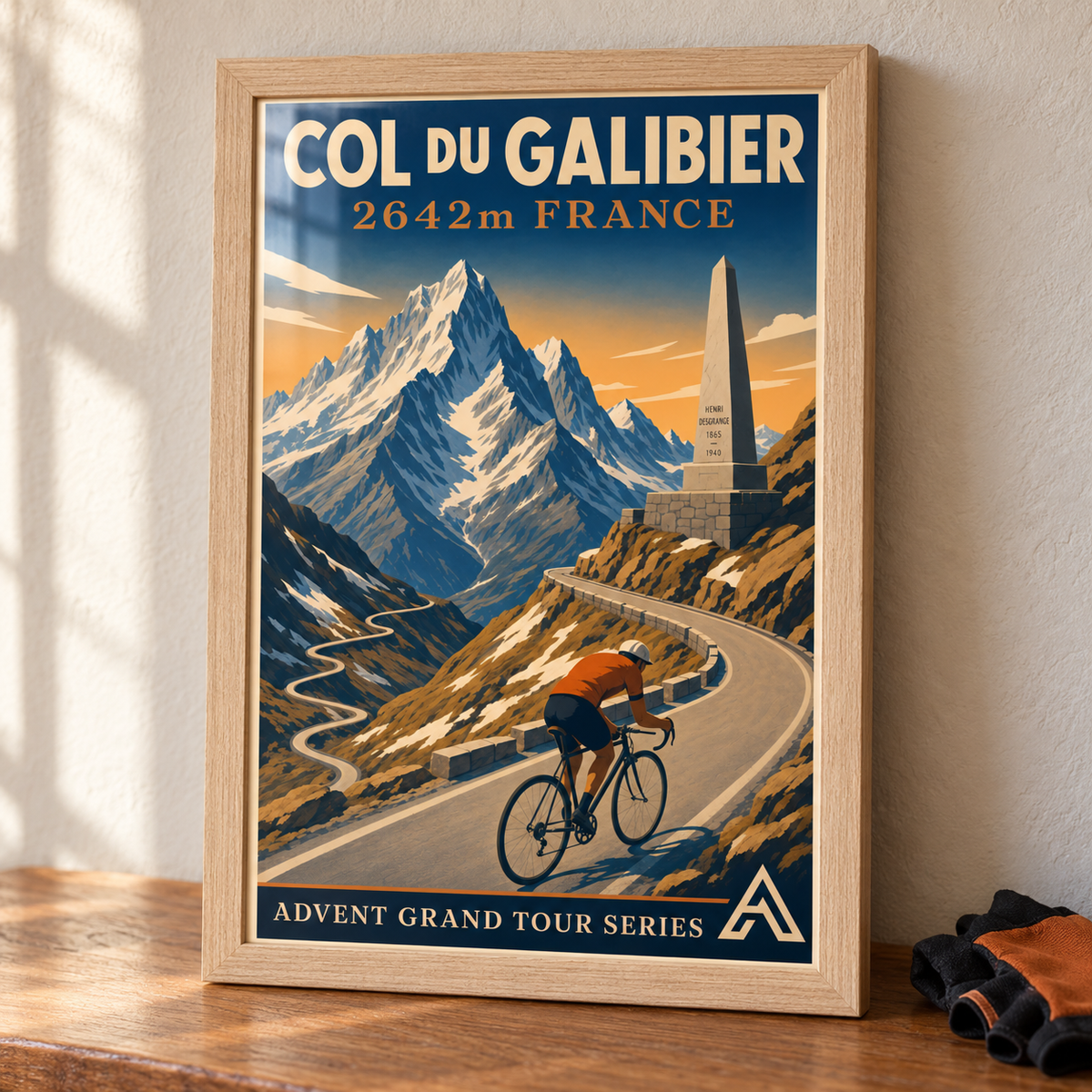 Col du Galibier art print in a Natural Oak frame displayed in full on an oak tabletop beside cycling gloves, showing a cyclist on an alpine mountain road.