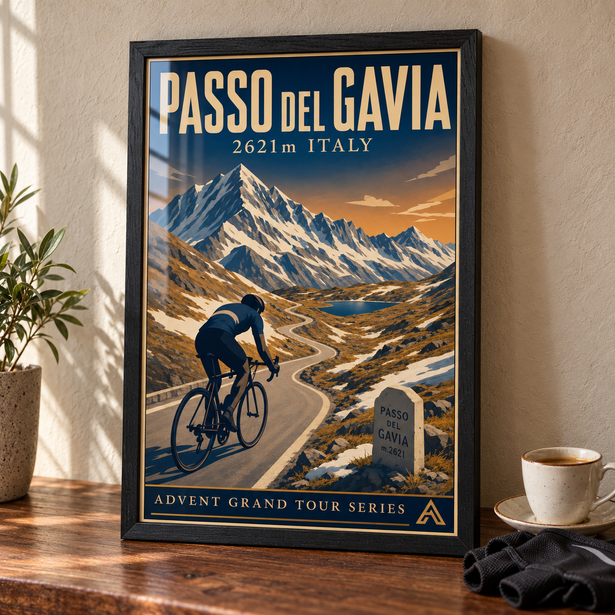 Passo del Gavia art print in a Black Ash frame on a wooden tabletop beside an espresso cup and cycling gloves, showing a cyclist, winding mountain road and alpine lake.