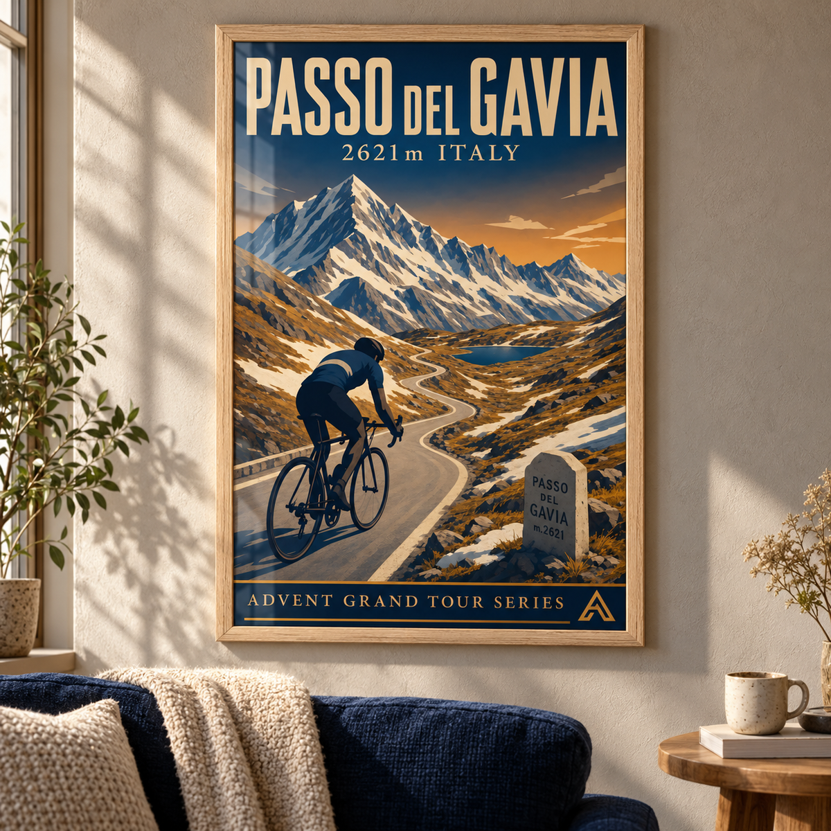 Passo del Gavia art print in a Natural Oak frame above a navy sofa in a sunlit mountain home, showing a cyclist on a winding road beneath snowy peaks.
