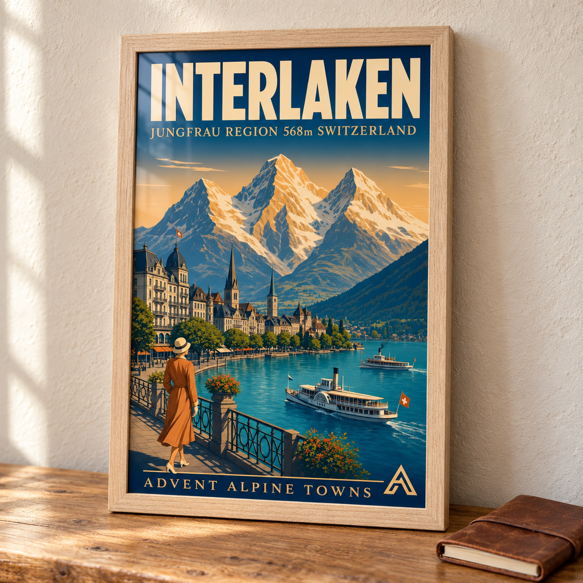 Interlaken art print in a Natural Oak frame on a wooden tabletop beside a leather travel journal, showing lakeside buildings, steamers and snowy Alpine peaks.