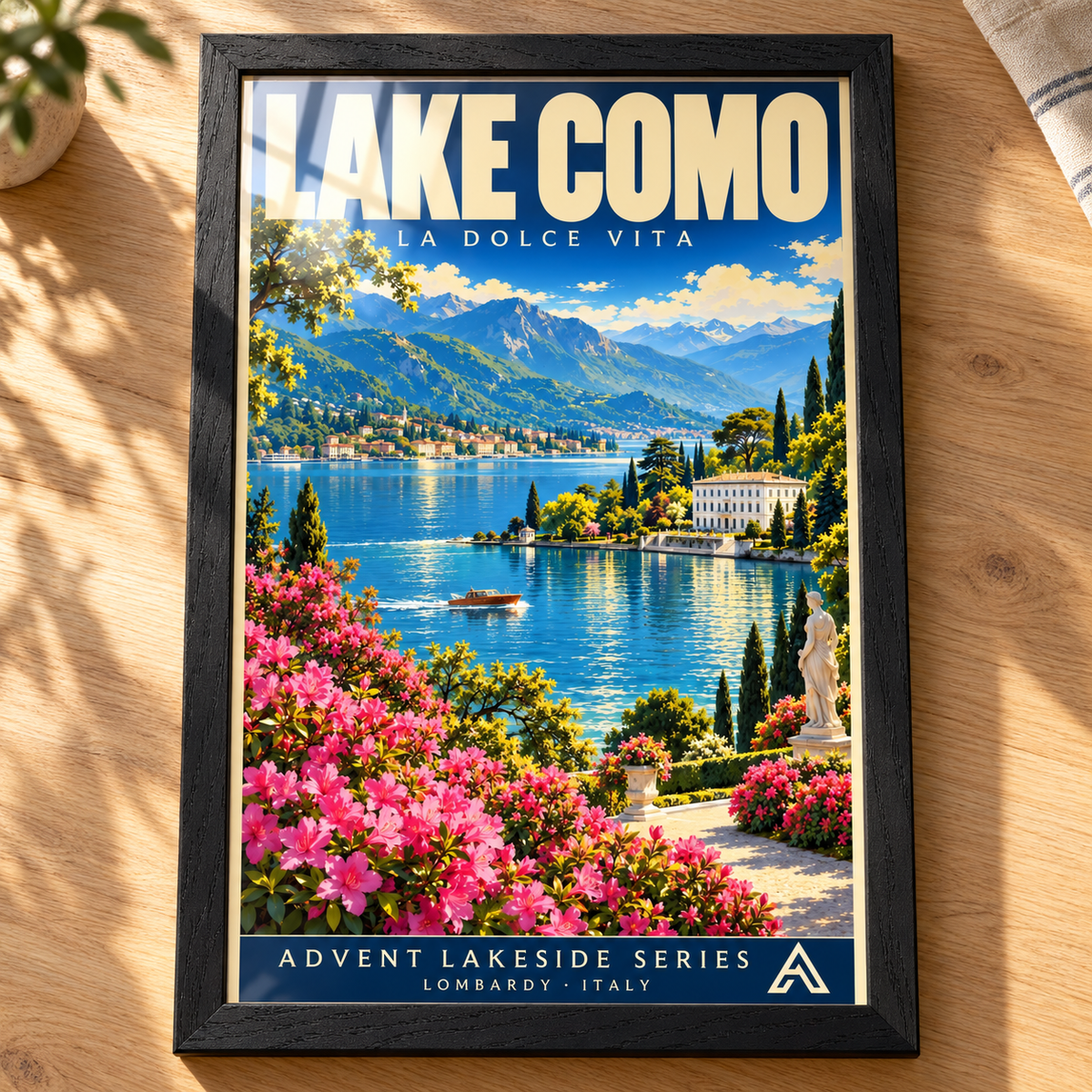 Complete Lake Como framed art print in Black Ash on an oak tabletop, showing a flower-lined terrace, blue lake, motorboat, villas and mountains, with sunlight reflecting across the glazing.