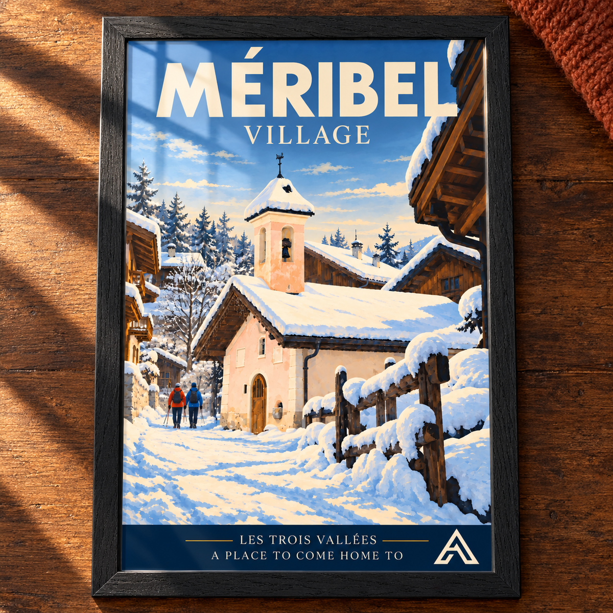 Complete Méribel Village art print in Black Ash on a dark oak tabletop, showing a snowy chapel, timber chalets and two walkers, with sunlight and window reflections on the glass.