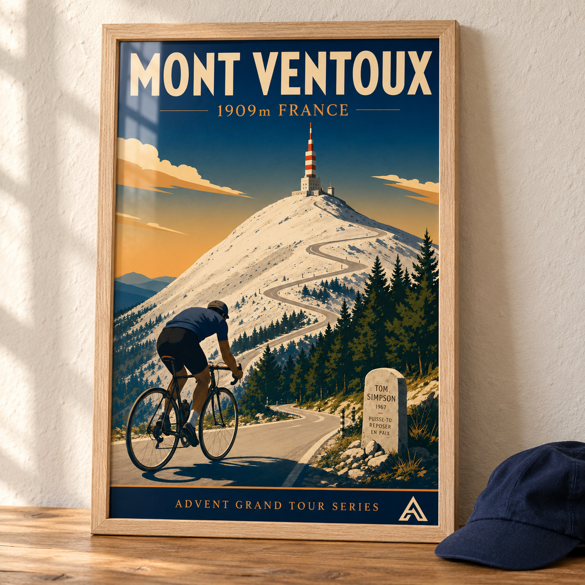 Mont Ventoux art print in a Natural Oak frame on a wooden tabletop beside a navy cycling cap, showing a cyclist on the winding summit road and the Tom Simpson memorial.