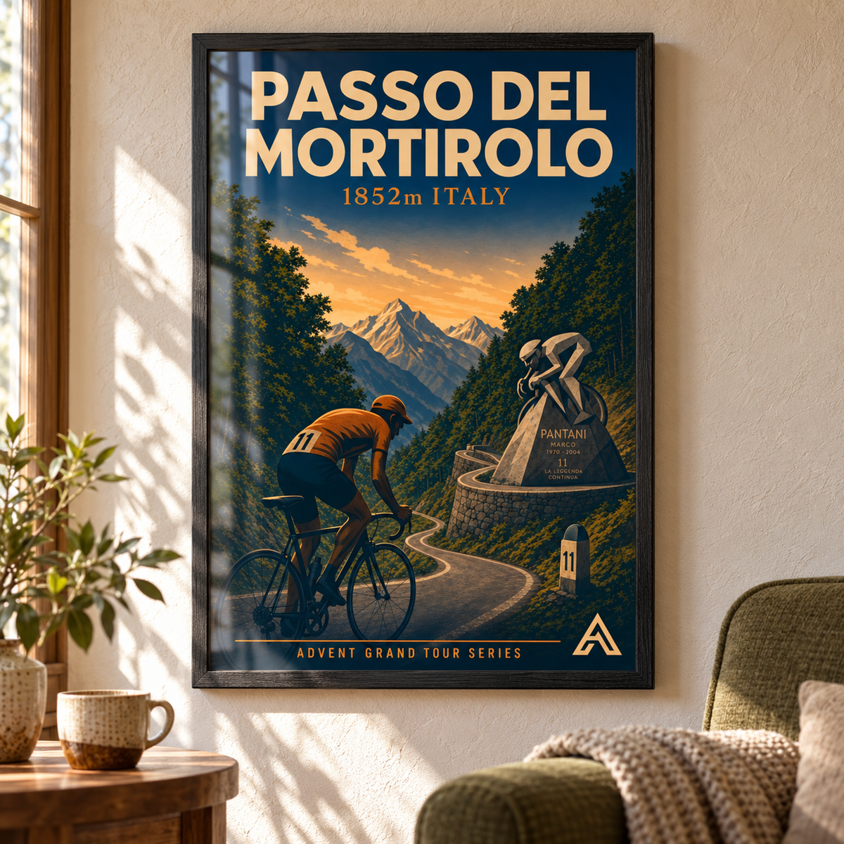 Passo del Mortirolo art print in a Black Ash frame beside a green armchair in a sunlit mountain home, showing a cyclist climbing past the Pantani memorial.