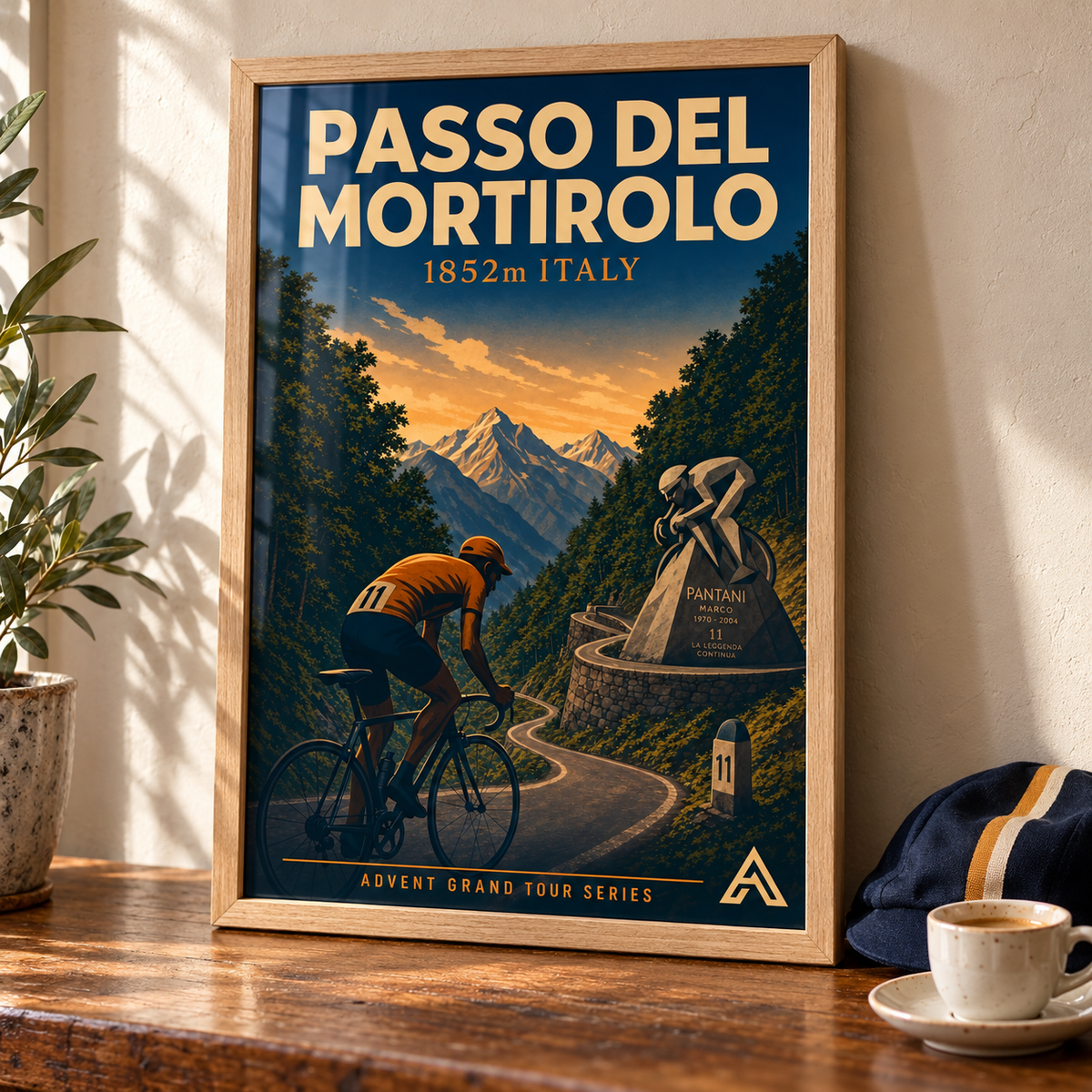 Passo del Mortirolo art print in a Natural Oak frame on a wooden tabletop beside an espresso cup and cycling cap, showing an orange-jersey cyclist on a forested mountain road.