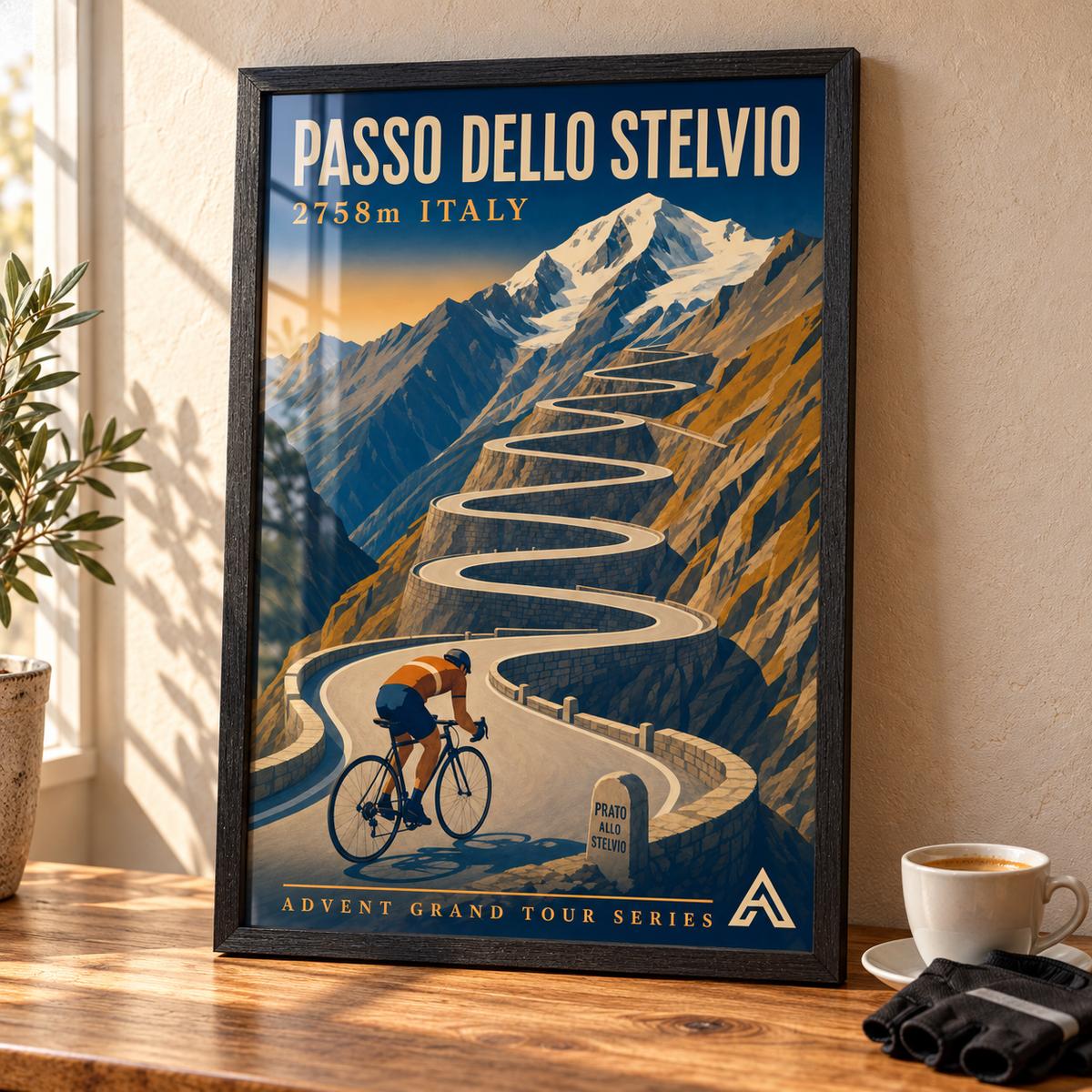 Passo dello Stelvio art print in a Black Ash frame on a wooden tabletop beside an espresso cup and cycling gloves, showing a cyclist climbing switchbacks beneath a snowy peak.